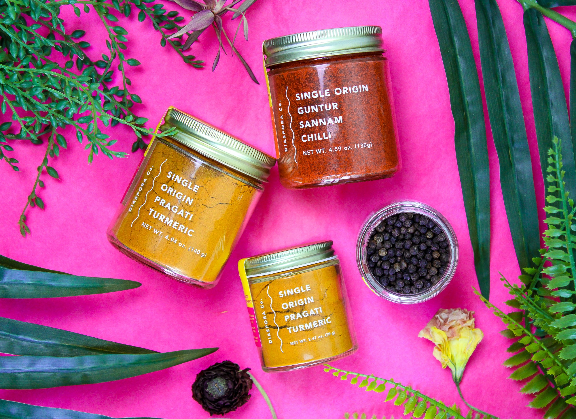 Jars of spices made by Diaspora Co against a pink background. One small jar of turmeric, one small open jar of peppercorns, one large jar of turmeric, and one large jar of Sannam chiles.